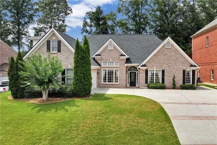 Traditional brick residence featuring a dark shingle roof, arched entryway, a concrete driveway, well-maintained lawn, and established landscaping