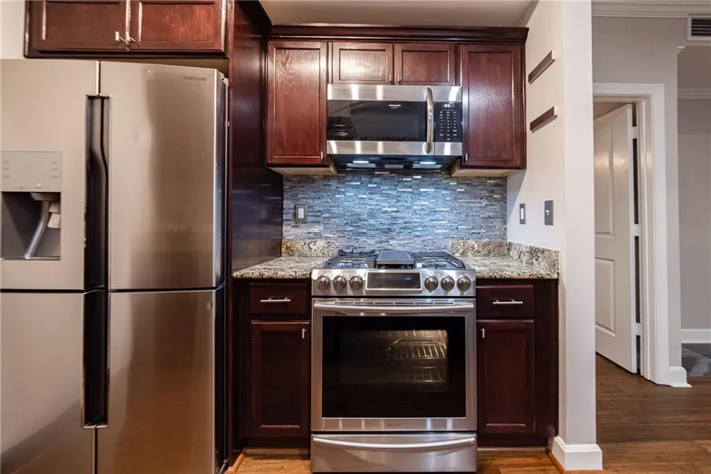 Kitchen with appliances with stainless steel finishes, wood finished floors, decorative backsplash, light stone countertops, and dark brown cabinetry
