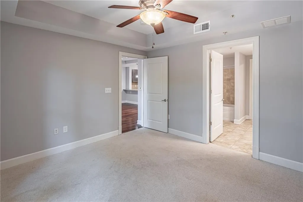 Unfurnished bedroom with ceiling fan, light colored carpet, a tray ceiling, and ensuite bath