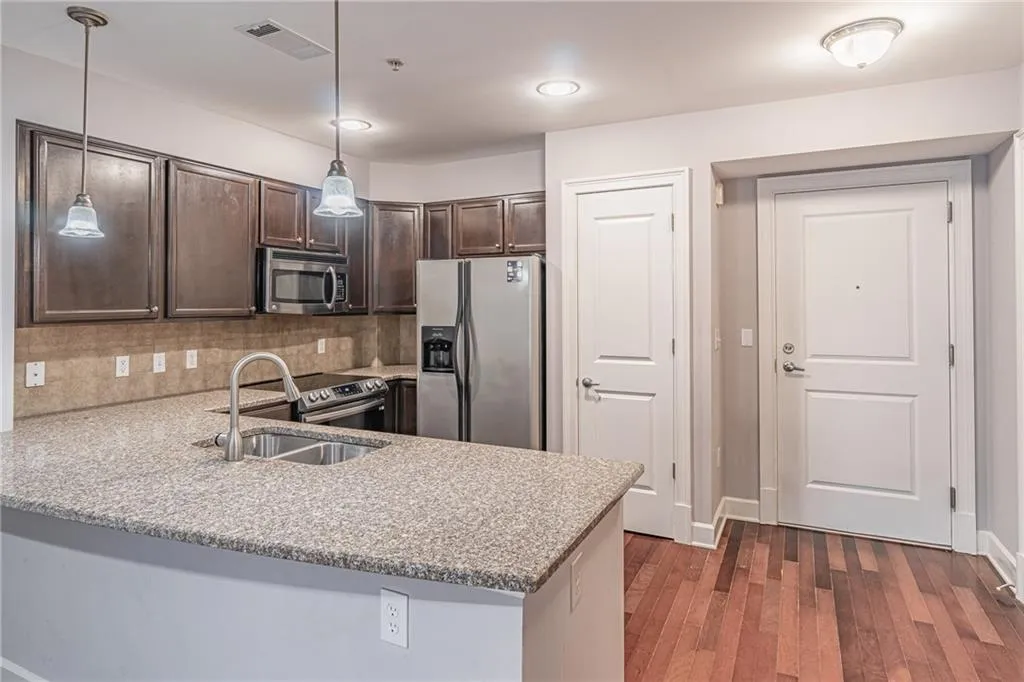 Kitchen with dark brown cabinetry, stainless steel appliances, hanging light fixtures, kitchen peninsula, and light stone counters