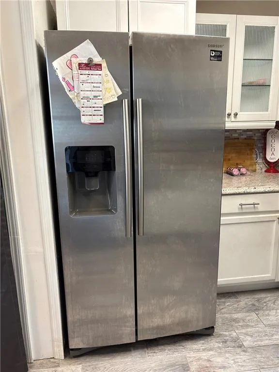 Kitchen view of stainless steel fridge with ice dispenser, white cabinetry, glass insert cabinets, and light stone countertops
