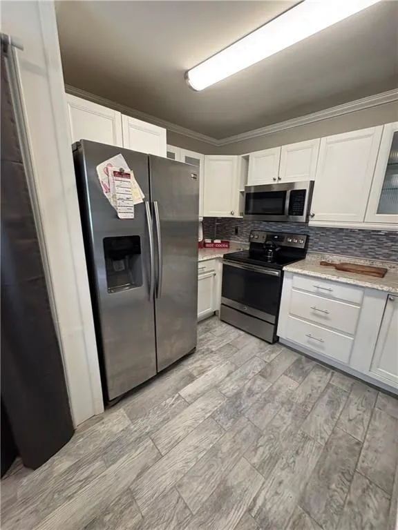 Kitchen featuring stainless steel appliances, ornamental molding, white cabinets, tasteful backsplash, and light stone countertops