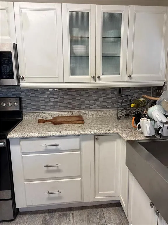 Kitchen featuring stainless steel microwave, black range with electric stovetop, white cabinetry, light stone countertops, and glass insert cabinets