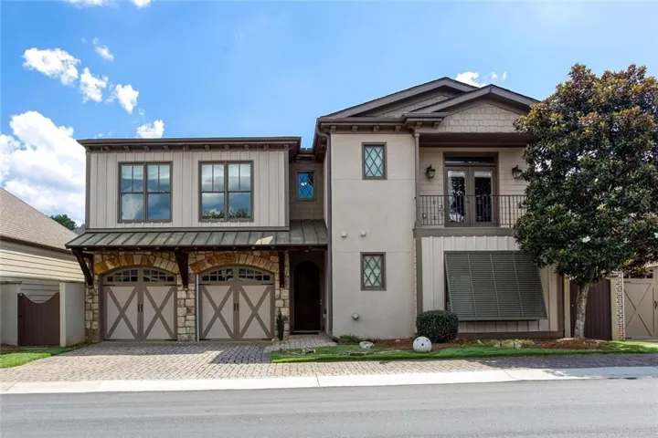 Multi-story facade featuring stucco and shingle siding, a two-car garage with stone archways, a metal roof accent, and a second-story balcony with metal railing