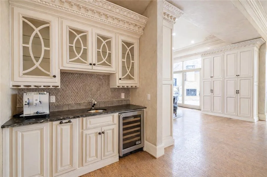 Indoor wet bar with wine cooler, dark stone countertops, crown molding, glass insert cabinets, and light wood-type flooring