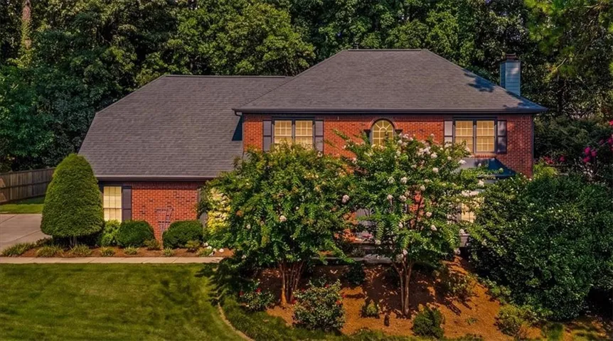 Brick exterior residence featuring a dark shingle roof, arched accent window, and mature landscaping