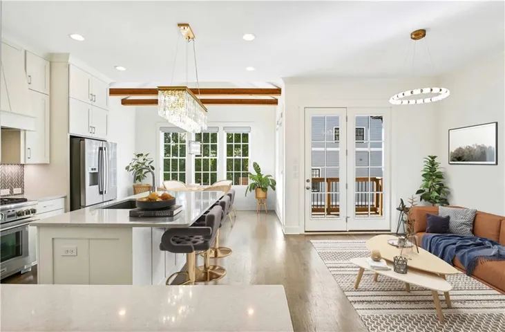 Open-concept living space with light wood-finish flooring, white cabinetry, a large kitchen island with counter seating, stainless steel appliances, and double French doors opening to an exterior deck