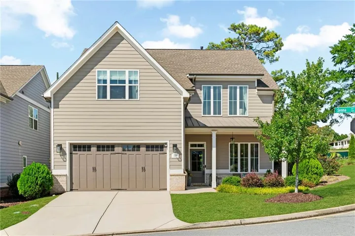 Craftsman style two story home in Marietta, Georgia with tan horizontal lap siding, white trim, and a standing seam metal roof accent over the covered front porch. Stacked stone skirting, an oversized carriage style two car garage, and manicured landscaping give the home strong curb appeal. Located on Senna Street adjacent to Marietta City Club Golf Course and just a few blocks from the historic Marietta Square.