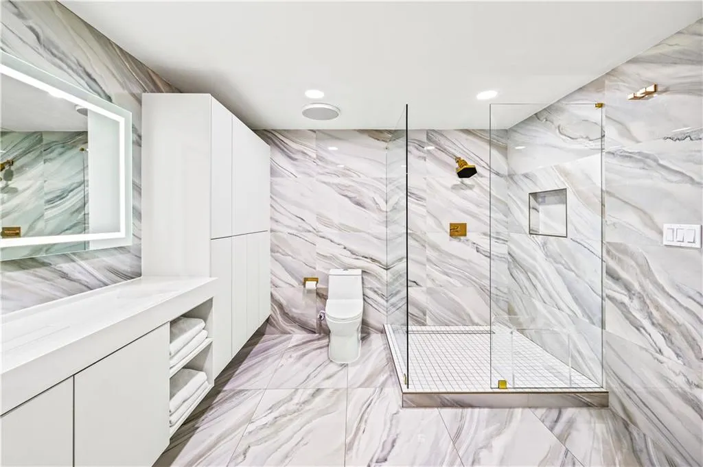 Bathroom featuring floor-to-ceiling stone-look tiling, white cabinetry, a glass-enclosed walk-in shower with gold-tone fixtures, and a wall-mounted illuminated vanity mirror