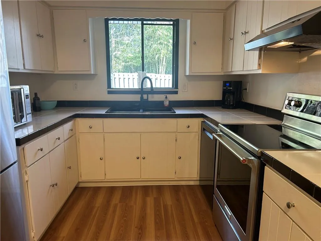 Kitchen with tile countertops, under cabinet range hood, wood finished floors, a sink, and electric stove
