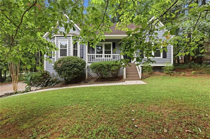 Charming cottage-style residence showcasing a manicured lawn with mature shade trees, a welcoming front porch with railings, and lush landscaping including boxwood shrubs