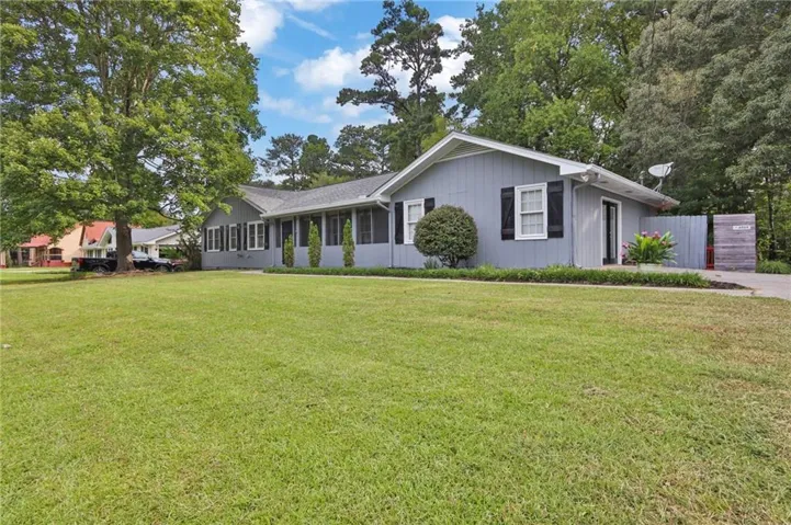Light blue siding exterior with dark shutters, covered front porch, a paved driveway, and mature trees framing the property