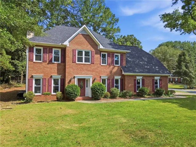 Brick exterior featuring multiple windows with shutters, a dark roof, a prominent entryway, and surrounding mature trees