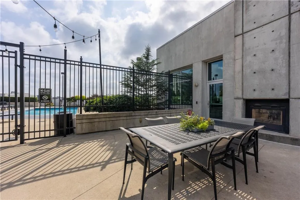 Outdoor patio with a built-in fireplace, concrete exterior, and a pool area beyond a black metal fence
