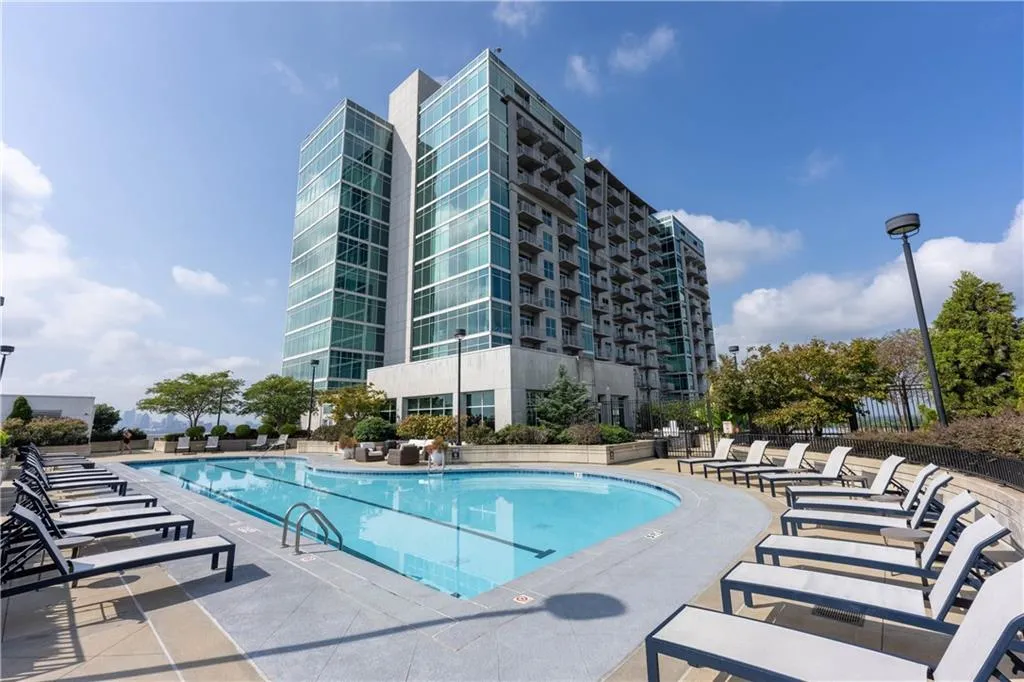 Outdoor swimming pool with integrated lap lanes, surrounded by a paved deck and lounge seating, set against a modern building with extensive glass facades and private balconies