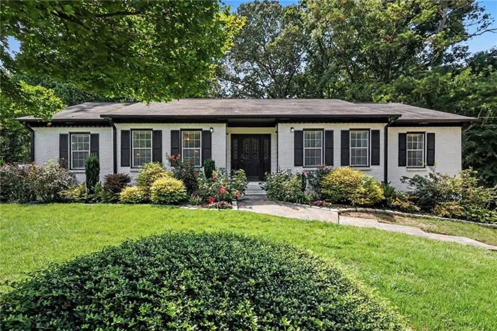 White brick exterior with dark shutters, established landscaping, front lawn, and a paved walkway
