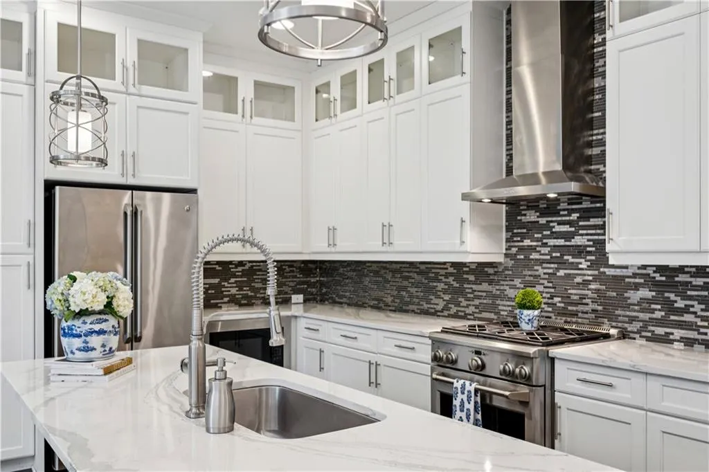 Kitchen with wall chimney exhaust hood, white cabinetry, stainless steel appliances, light stone counters, and hanging light fixtures