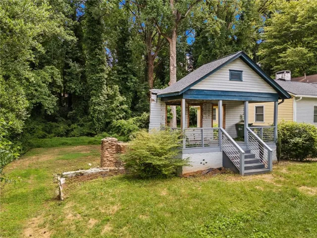 Gabled roofline with light siding and blue trim accents, front porch with wood columns and metal railings, exterior stairs, stone wall feature, and abundant surrounding greenery