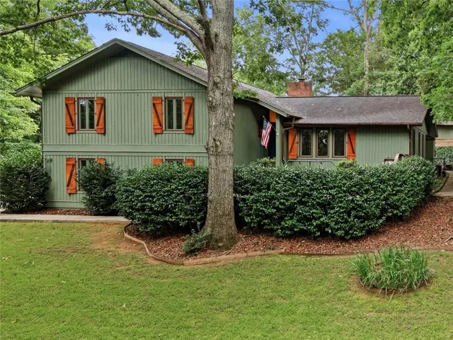Sage green vertical siding exterior featuring contrasting rustic orange shutters, a mature front yard tree, established shrubbery, and a well-maintained lawn