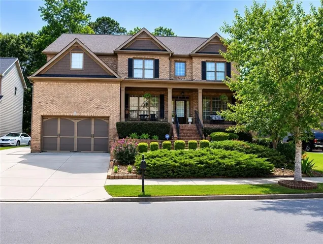 Brick facade residence featuring a two-car garage with arched doors, a covered front porch with dark railings, a prominent front entry, and mature landscaping