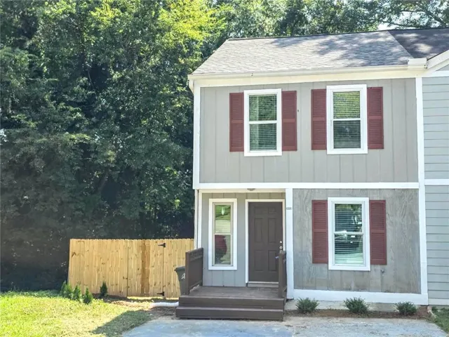 Two-story exterior with light gray siding, brown shutters, a dark brown entry door, a front porch with dark brown railings, and a wood privacy fence