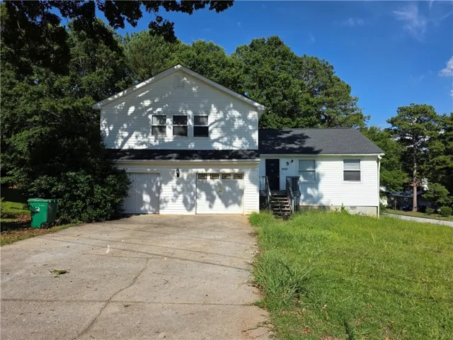 White siding exterior with a two-car garage, dark shingle roof, and mature perimeter trees