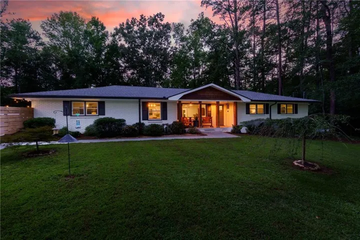 White brick exterior with dark shutters, covered front porch, prominent wood pillars, wood-finish front door, and mature surrounding trees