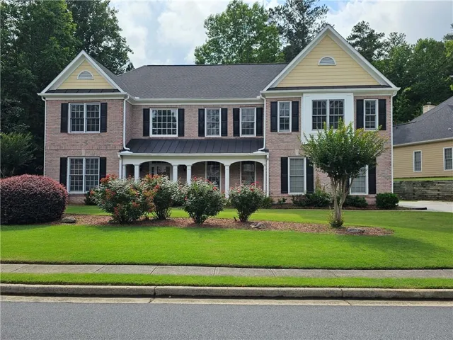 Traditional brick-front residence featuring a covered porch with white columns, dark shutters, and a well-maintained front lawn with established plantings