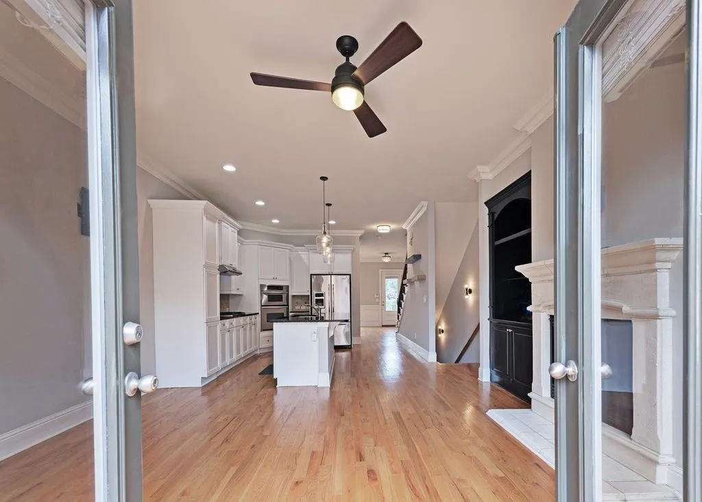 Open Plan Living/Family Room And Kitchen Area Featuring Built In Bookcases, Fireplace, Crown Molding, Ceiling Fan, and Light Hardwood Floors.