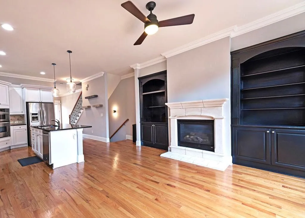 Open Plan Living/Family Room And Kitchen Area Featuring Built In Bookcases, Fireplace, Crown Molding, Ceiling Fan, and Light Hardwood Floors.