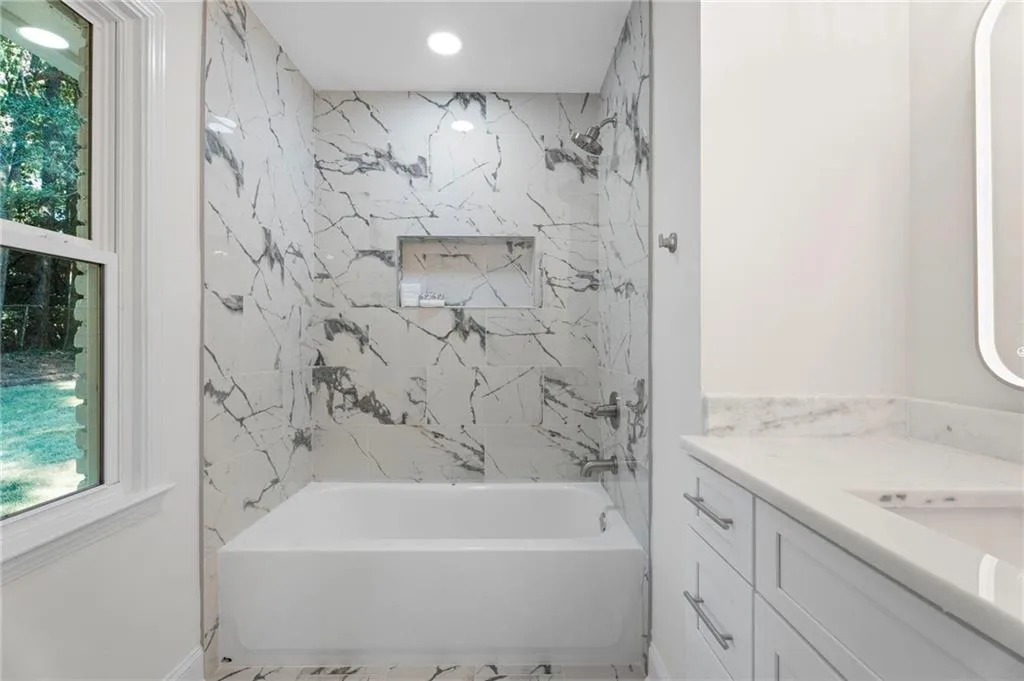 Spa-inspired primary bathroom featuring marble-look tile surround with veining detail, a soaking tub with integrated shower enclosure, chrome fixtures, recessed ceiling lighting, a floating vanity with dual drawers, and a large mirror with integrated lighting