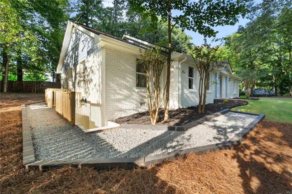 White painted brick exterior with clapboard siding, mature trees, landscaped beds with dark mulch, gravel pathways with dark wood borders, and a private wooden fence