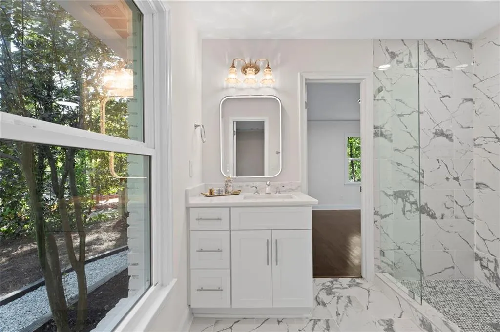 Spacious bathroom featuring a white vanity with integrated sink, rectangular illuminated mirror, glass-enclosed shower, and large format marble-look tile flooring