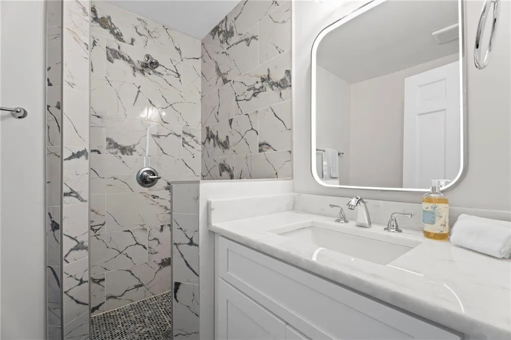 Walk-in shower with tiled walls and floor, vanity with white cabinetry, white countertop, and integrated sink, and an illuminated mirror