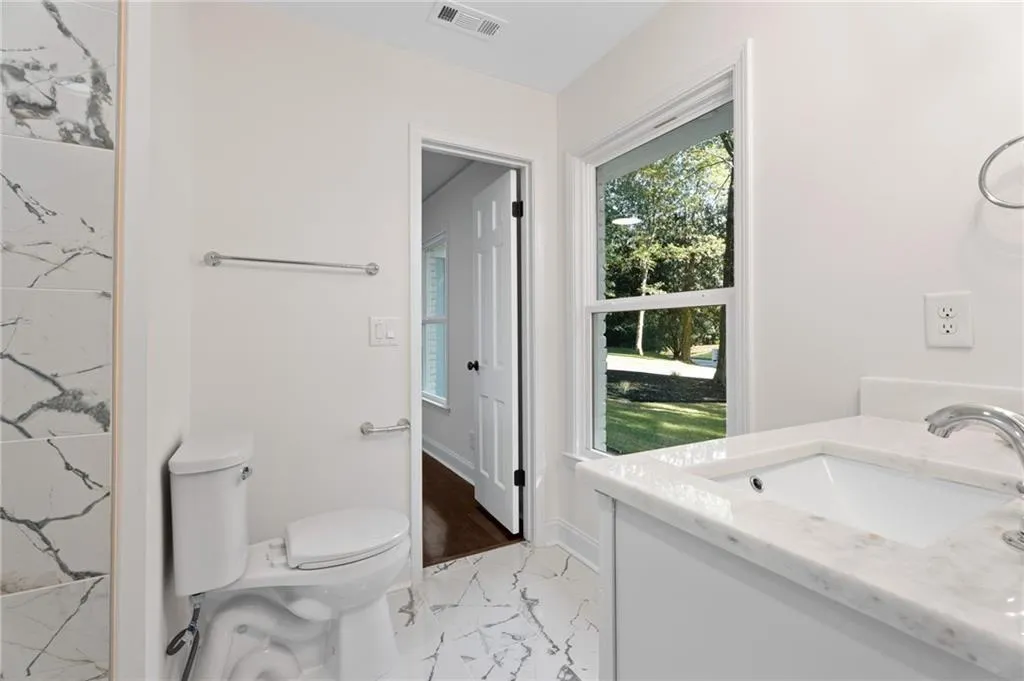 Spa-inspired primary bathroom featuring marble-look tile flooring, a white subway tile shower enclosure with frameless glass partition, white painted cabinetry, a marble-look vanity countertop with integrated sink, and abundant natural light from an oversized window framing manicured landscaping and mature trees