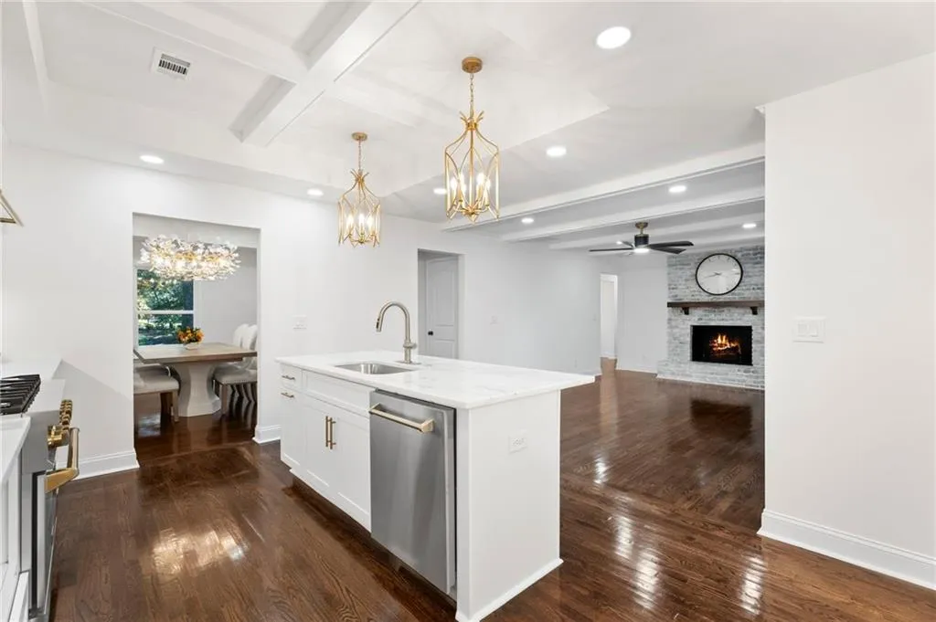Contemporary kitchen and great room featuring white cabinetry with brushed gold hardware, a waterfall-edge island with integrated sink and stainless steel appliances, warm wood-finish flooring throughout, recessed and pendant lighting, and an adjacent fireplace with dark surround
