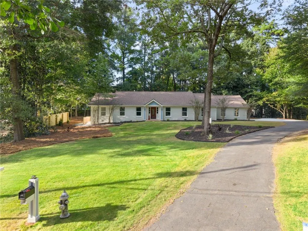 Ranch-style residence featuring a prominent driveway, mature trees, manicured lawn, and a light-colored exterior with a contrasting front door