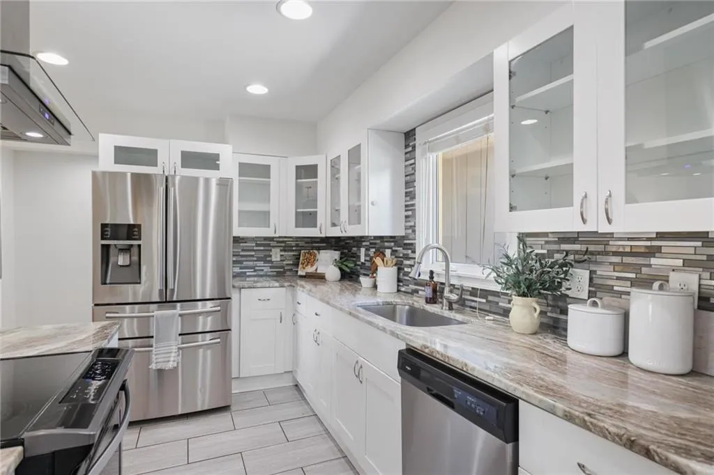 Kitchen featuring white cabinetry, appliances with stainless steel finishes, glass insert cabinets, extractor fan, and light stone counters Kitchen featuring white cabinetry, appliances with stainless steel finishes, glass insert cabinets, extractor fan, and light stone counters