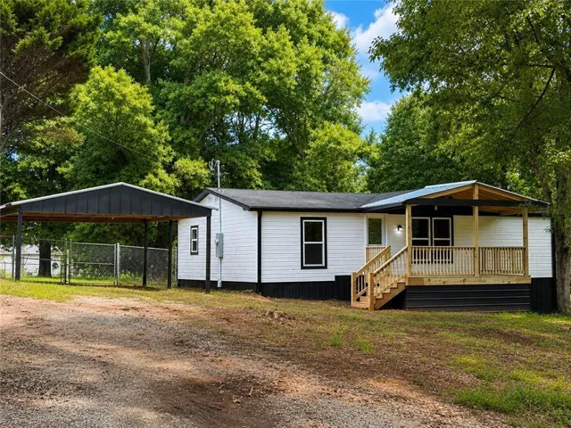 White exterior featuring a covered front porch with natural wood railings, a gravel driveway, and a black metal carport