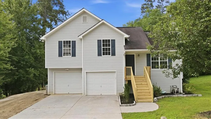 Split-level residence featuring light gray siding with dark blue window shutters, a two-car garage, a concrete driveway, and a natural wood staircase leading to the main entry