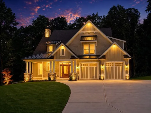 Craftsman-style residence featuring a prominent stone chimney, a covered porch with stone-clad columns, a wood-finish entry door, and a two-car garage with panel-style doors. Digitally Enhanced Twilight Image