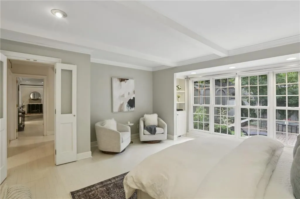 Beamed ceilings, white hardwood flooring, built-ins, French doors and encased wall of windows create a serene space and delightful sitting area in the primary suite. Private view of nature makes this a delightful space to unwind.
