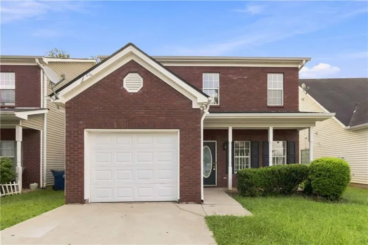 Brick facade with white trim, covered front porch, attached garage with white door, concrete driveway, and manicured shrubs