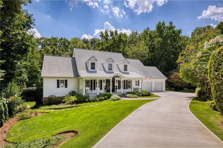 White clapboard siding with black shutters, dormer windows, a covered front porch, an attached two-car garage, and a curved concrete driveway