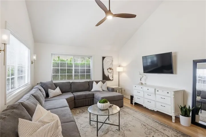 Vaulted ceilings enhance this living space, featuring wood-finish flooring, a contemporary ceiling fan, and multiple windows with horizontal blinds