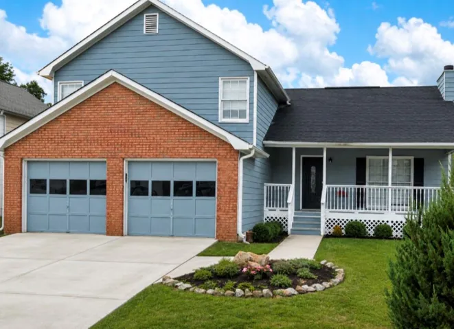 Exterior featuring brick and horizontal siding, a two-car garage with panel doors, a covered front porch with white railings, a concrete driveway, and a landscaped front yard with mature greenery