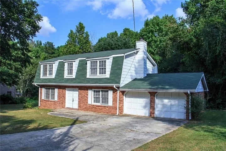 Dormered gambrel roofline with green shingles, brick exterior, white paneled double-door entry, double-car garage, and mature surrounding trees