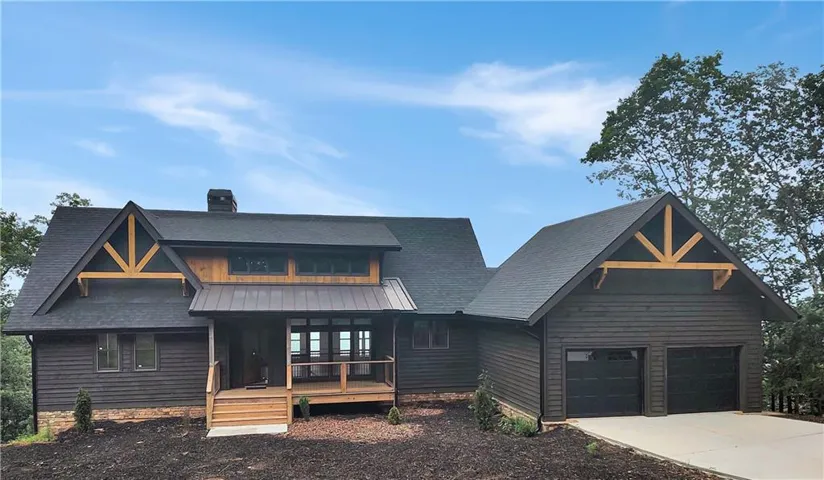 Modern farmhouse design featuring a dramatic timber-frame gable exterior with mixed dark siding and warm wood accents, expansive front-facing windows with multi-pane detailing, a welcoming covered entry porch with wood decking, and a professionally finished two-car garage with matching architectural elements