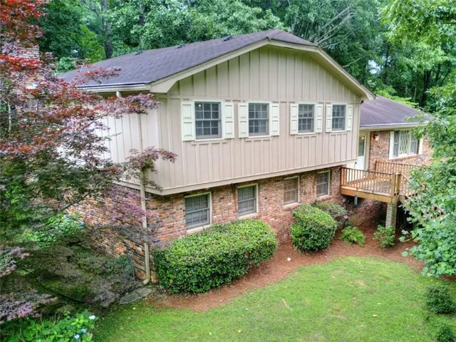 4-level home featuring brick and vertical siding, a wooden deck, and a sloped lawn with mature landscaping.