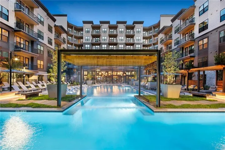 Resort-style courtyard featuring an illuminated lap pool with integrated spa jets, metal pergola with overhead lighting, and multi-story residential buildings surrounding the amenity space